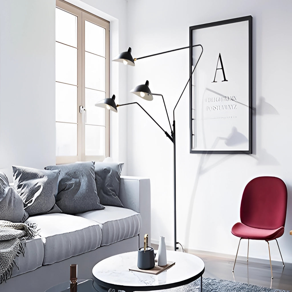 A floor lamp with a tripod base and directional light next to a white couch in a modern living room with large windows, black accents, and picture frames on the wall.
