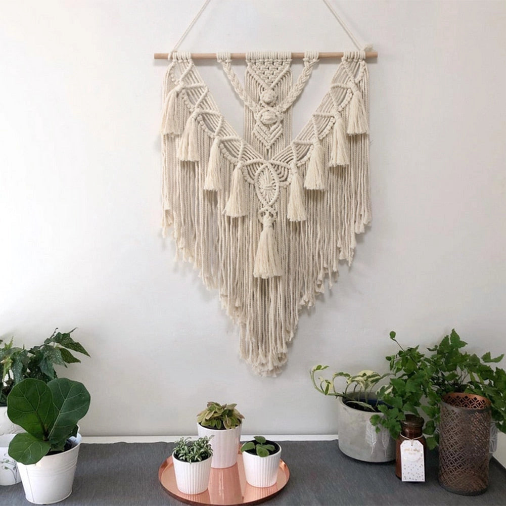 Handwoven macrame boho wall hanging tapestry with geometric design, displayed on a white wall, accompanied by houseplants and a flowerpot in a stylish interior setting.