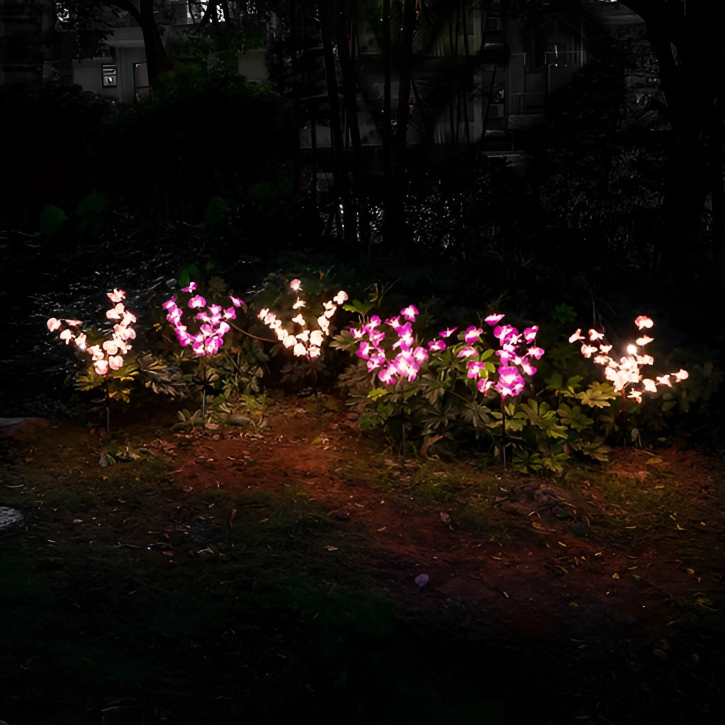 Twin pack of LED solar orchid lights with color transition placed in a natural environment, featuring realistic flowers with petals and branches among grass and groundcover.