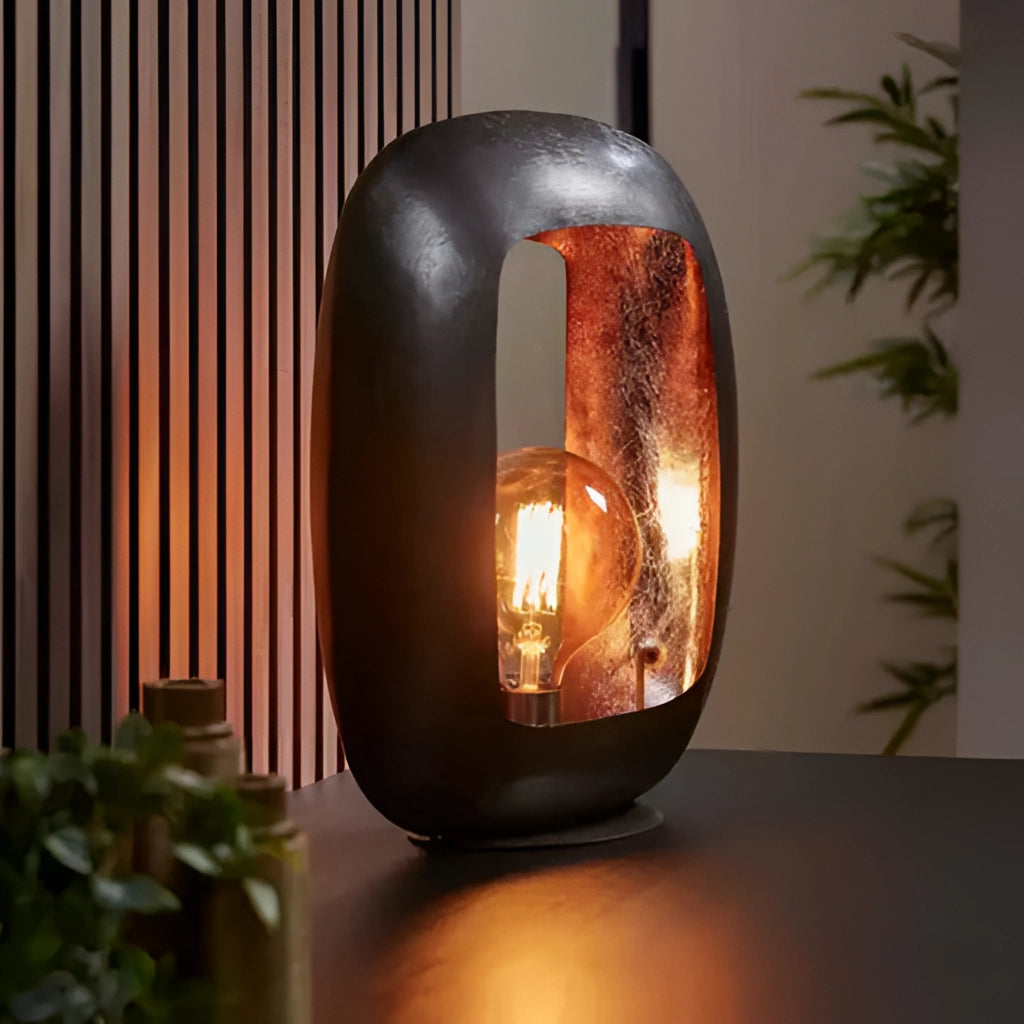 Large metal crafted vintage style table lamp with an amber glass shade, placed on a wooden surface with a small plant nearby.