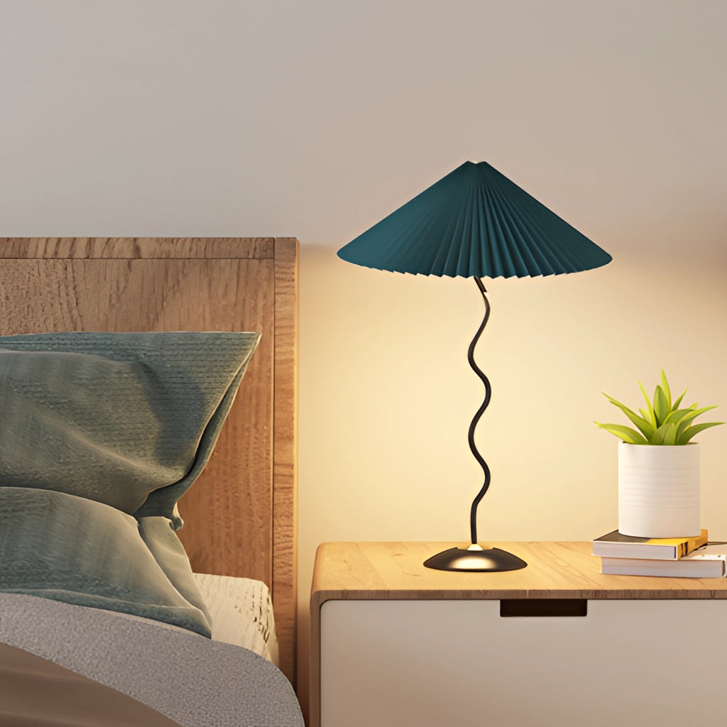 A curved pleated floor lamp with a metal textile shade, placed in a well-designed interior setting featuring wooden furniture, a potted plant, and ambient lighting.