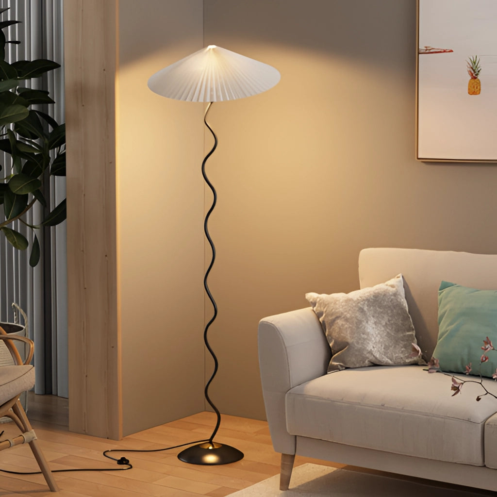 A modern metal textile floor lamp with a curved, pleated white shade, wooden base, and standing on a hardwood floor in a well-lit, comfortable interior setting.