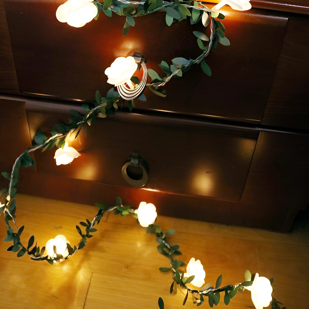 Battery-operated rose flower string lights with LED garland, featuring green leaves and amber lights, adorning a wooden interior wall.