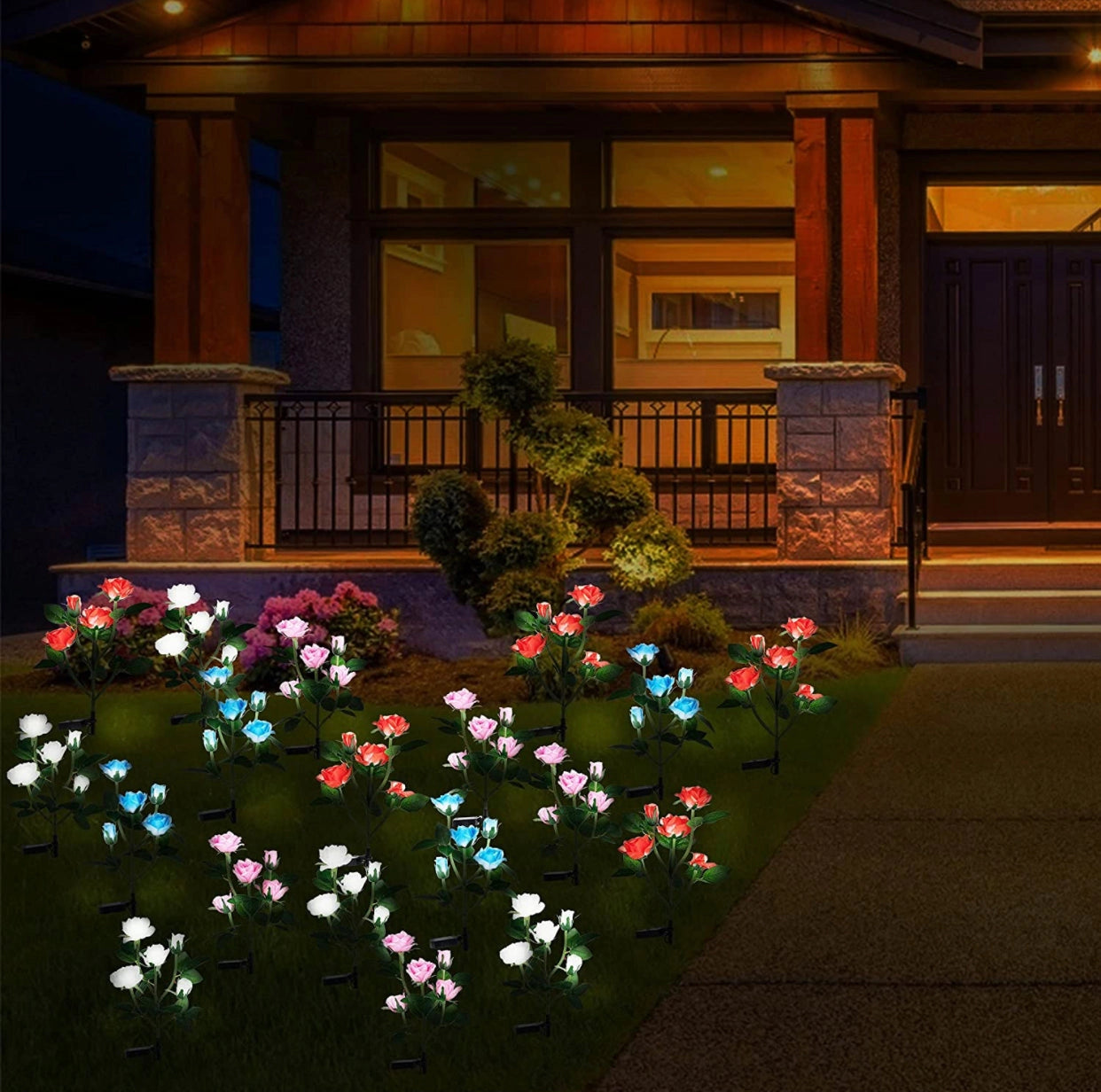 Solar LED Rose Lights Twin Pack illuminating a garden area at night, featuring white rose-shaped lights placed among plants near a residential building.