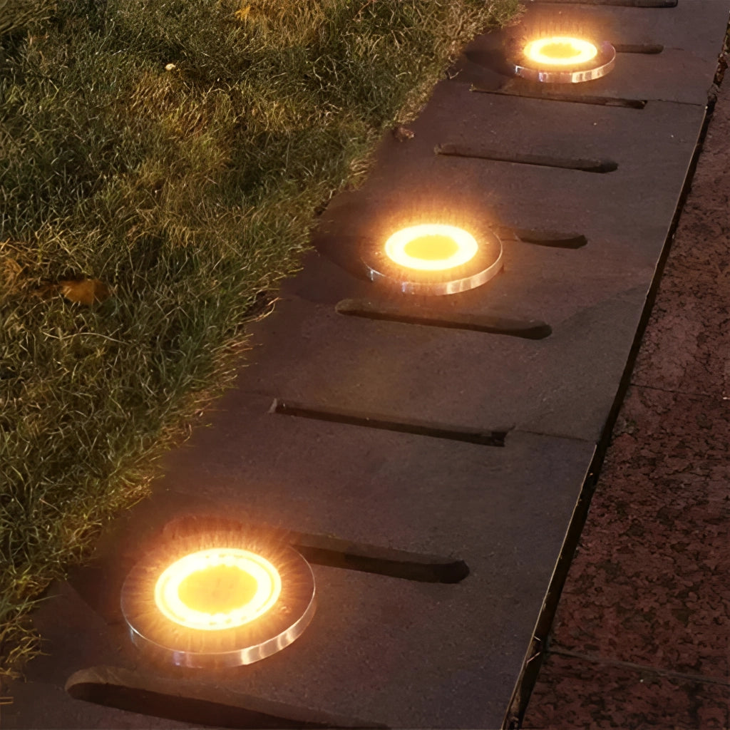 Solar pathway disk lights illuminating a garden pathway at dusk with surrounding grass and asphalt surface.