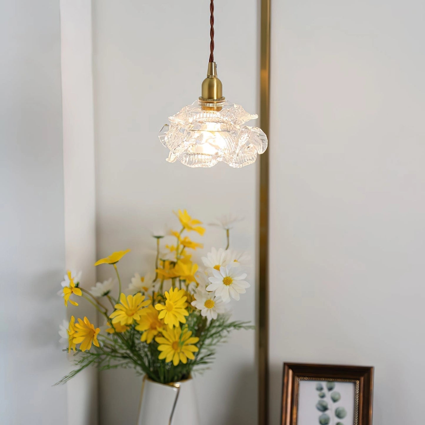 Vintage brass wall light with rose glass shade, mounted on a wall, elegantly illuminating a room decorated with a flower in a vase, picture frames, and wooden accents.