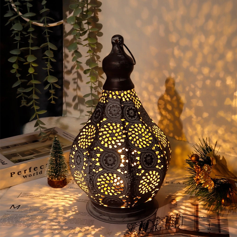 Vintage Moroccan metal lantern with intricate designs, battery-powered, placed among plants and ornaments, featuring tints and shades of metal and glass, suitable for events and artistic decor.