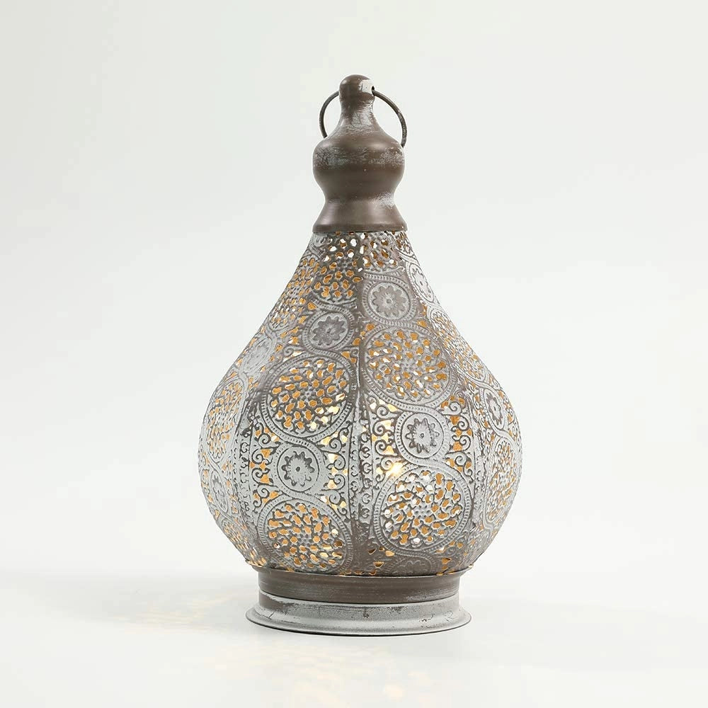 Vintage Moroccan Metal Lantern with intricate design, battery-powered, displayed on a wooden surface with artistic elements in the background.