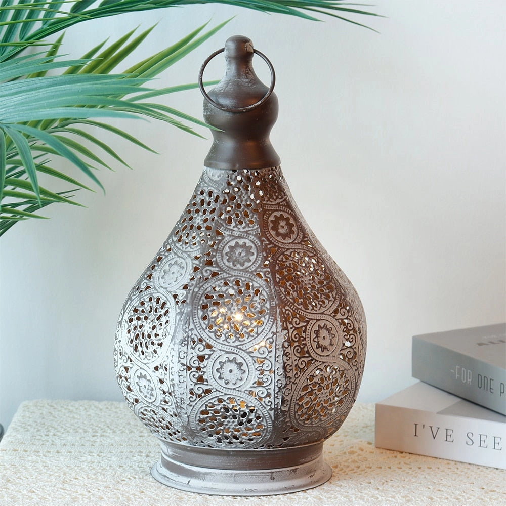 A vintage Moroccan metal lantern with intricate designs, placed on a wooden table next to a small plant and pottery, with a window in the background.