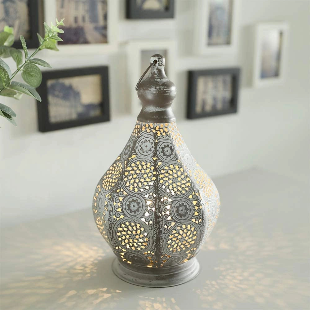 A vintage Moroccan metal lantern with intricate designs, illuminated by battery power, placed on a wooden floor near a window with interior design elements including picture frames, plants, and artwork.