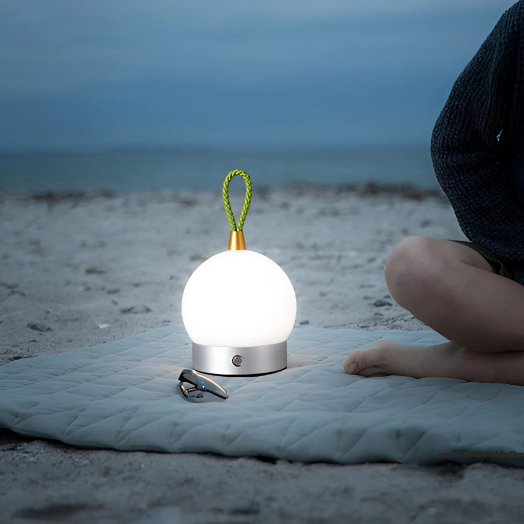 Waterproof LED cordless table lamp with orb design, illuminated by sunlight, positioned near a beach with a clear blue sky and calm body of water in the background.
