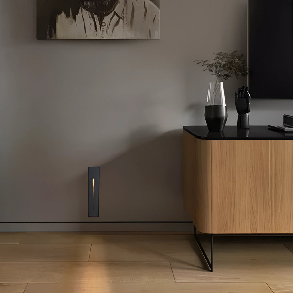 Modern ambient glow wall light in black, white, and light grey, mounted on a sleek indoor wall, surrounded by minimalist decor including a picture frame and a plant, set against a grey hardwood floor.