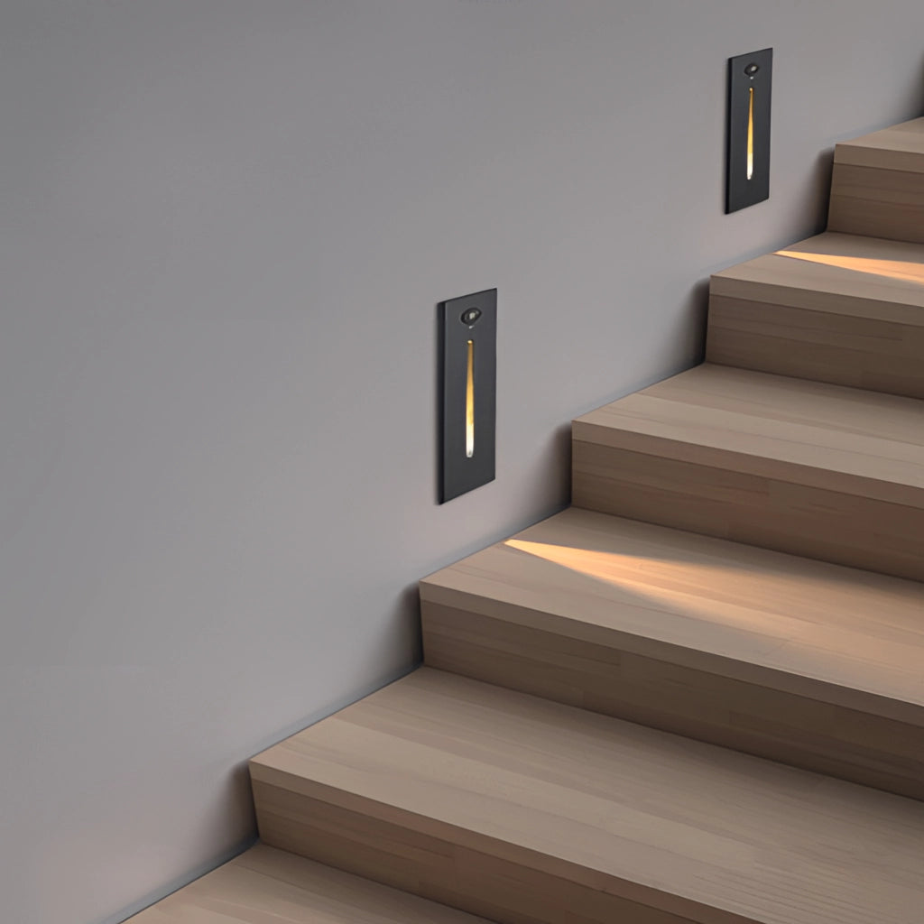 Modern stylish ambient glow wall light in black, white, and light grey colors, mounted on a wooden wall. The rectangular aluminum light fixture is suitable for both indoor and outdoor use, with waterproof IP65 rating. The surrounding area includes grey flooring and wooden stairs.