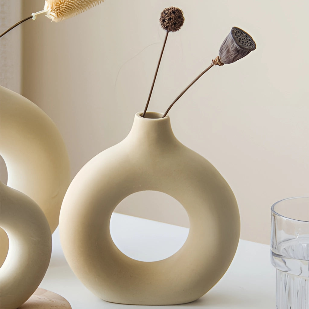 A ceramic donut-shaped vase with a modern design, featuring a minimalist aesthetic and holding a small twig. The vase is illuminated by soft lighting that highlights its smooth texture and elegant curves.