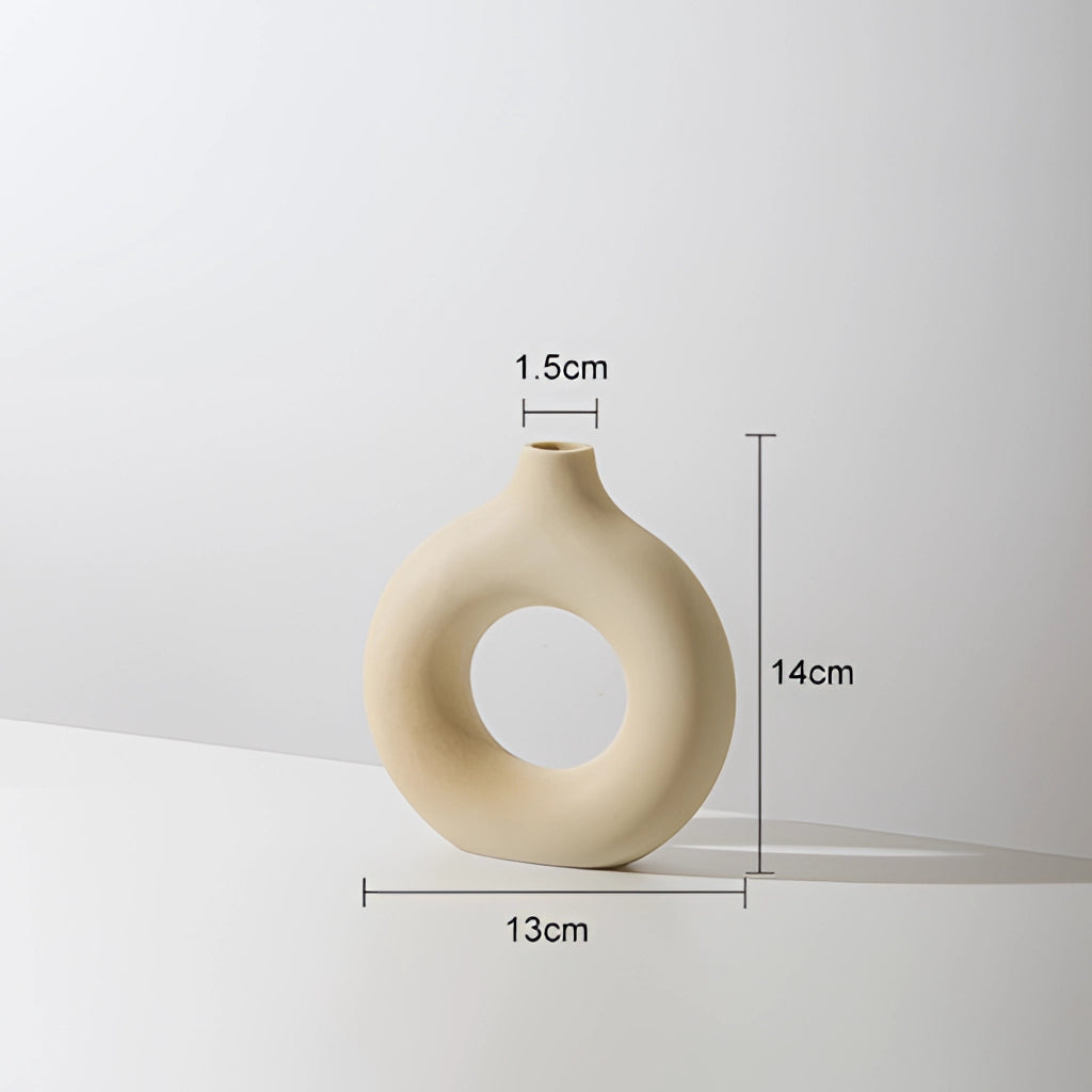 Beige ceramic donut-shaped vase for modern home decor placed on a wooden surface with a ceiling lamp in the background.