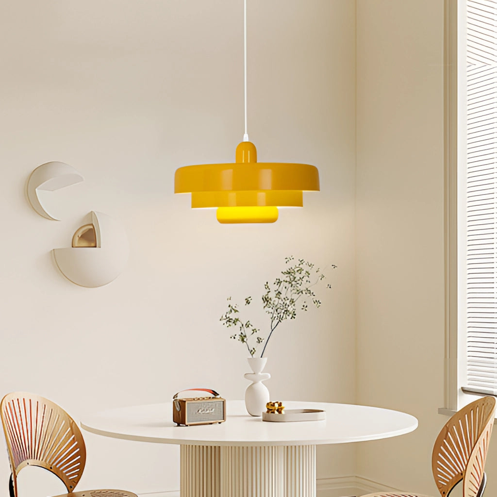 Modern metal pendant lamp with linked circles in yellow, displayed in a contemporary interior with a wooden table, white furniture, and a plant in the background.