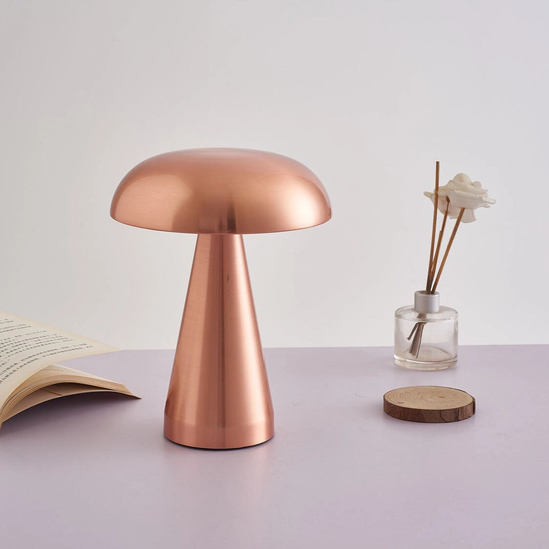 Cordless Mushroom LED Table Lamp in Rose Gold with Steepless Dimming sits on a wooden table next to a chair.