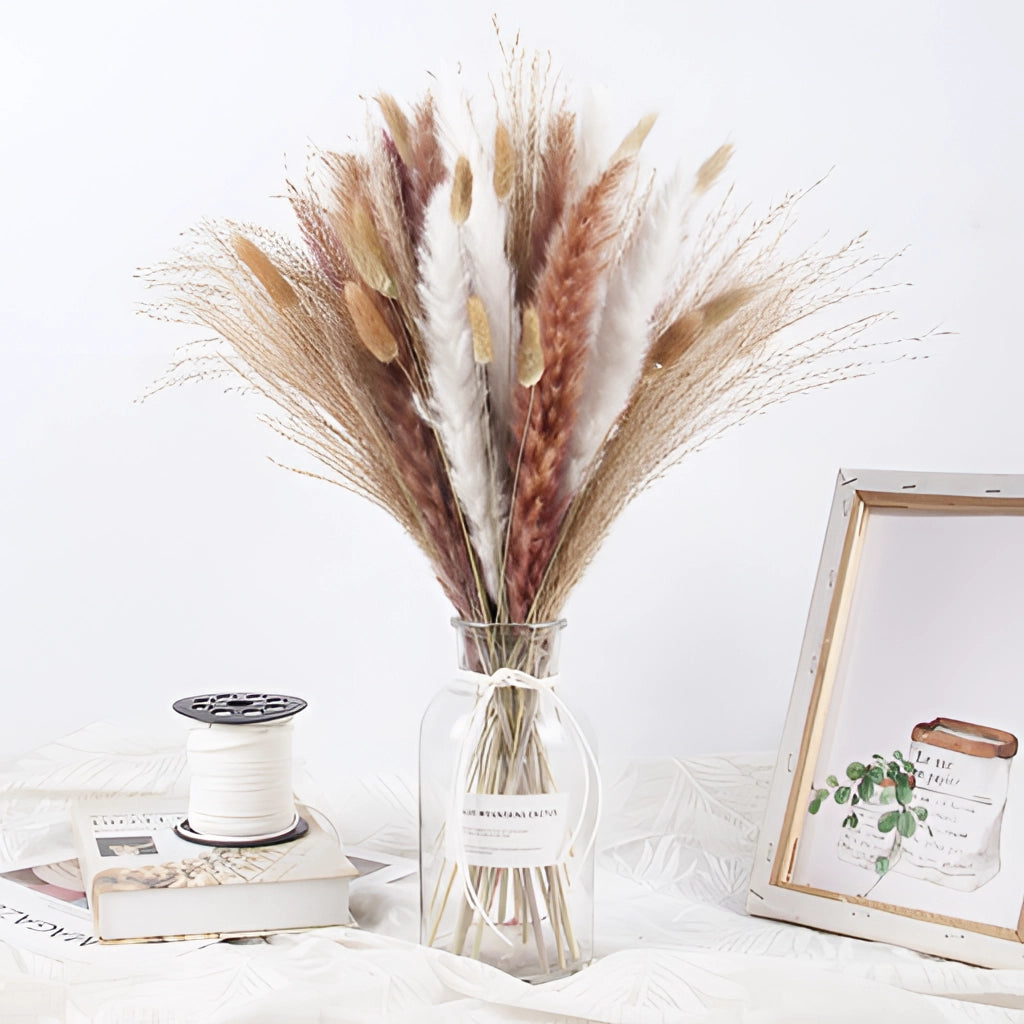 A set of dried pampas grass flowers arranged in a vase, placed on a table next to a picture frame and serveware, creating a natural and artistic interior decoration.