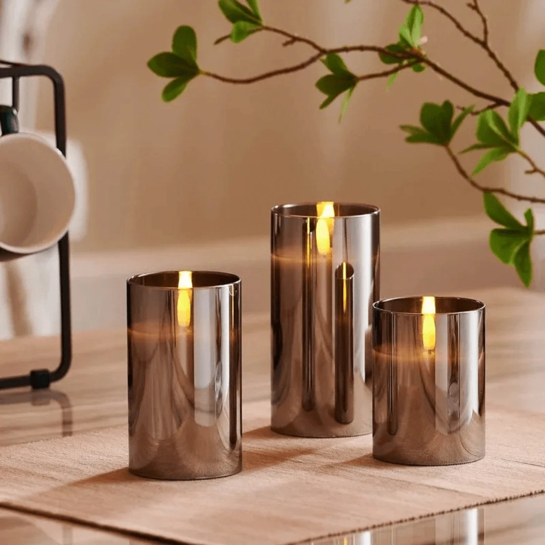 Flameless glass candle set with remote control, featuring three LED pillar candles on a wooden surface, surrounded by plants and modern interior decor.