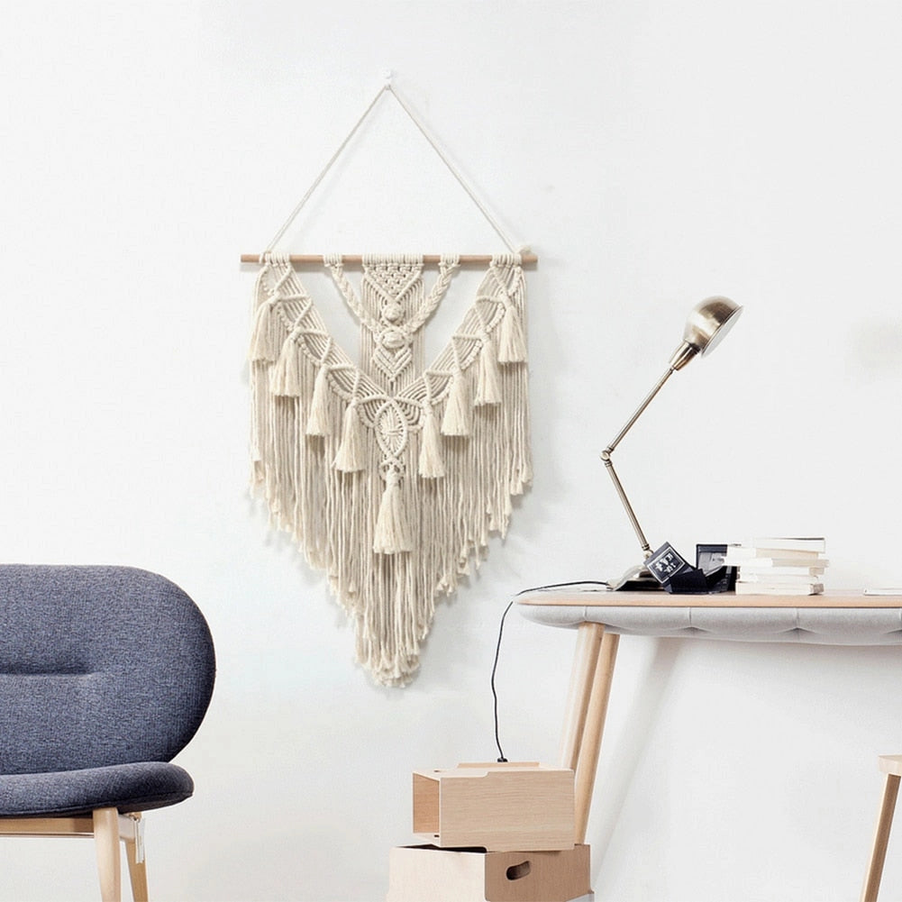Handwoven macrame boho wall hanging tapestry featuring geometric art design, displayed on a grey wall with wooden furniture and shelving in the background.