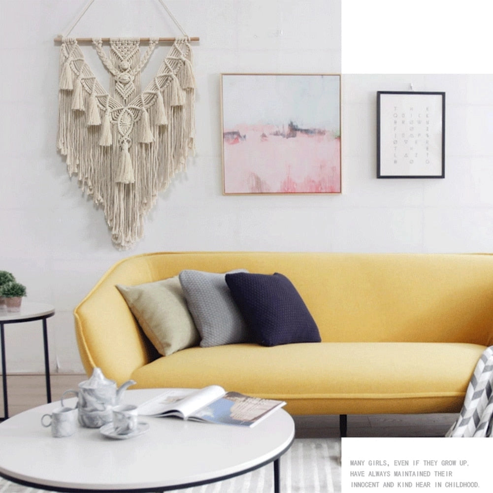 Handwoven macrame boho wall hanging tapestry with geometric art decor displayed above a white couch with a small table and a plant nearby.