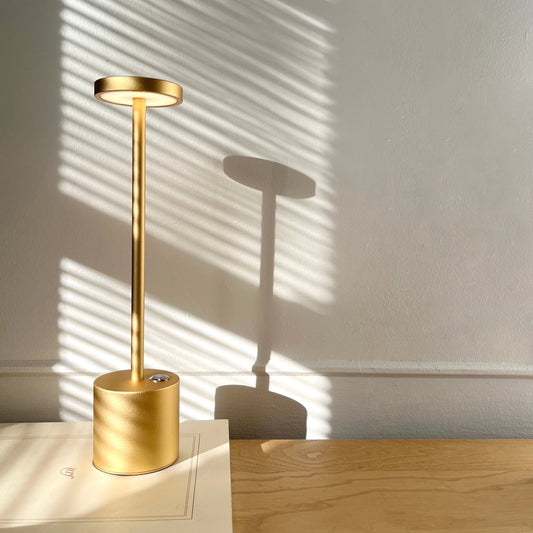 LED Portable Table Lamp with Resilient Design in Gold on a wooden surface with a wooden wall in the background.