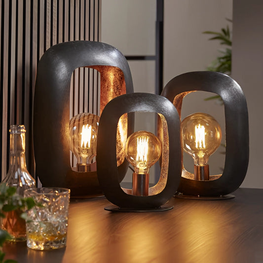 Vintage style metal crafted table lamp on a wooden surface with interior design elements in the background, including glass tableware and barware.