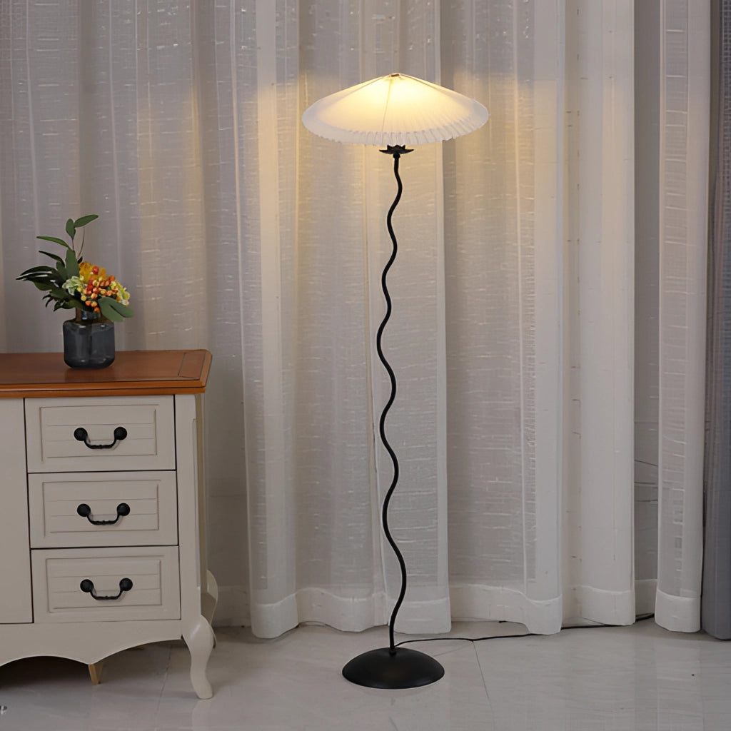 A metal textile curved pleated floor lamp stands beside a wooden cabinet with drawers, situated in a well-designed interior space with grey walls and a potted plant.