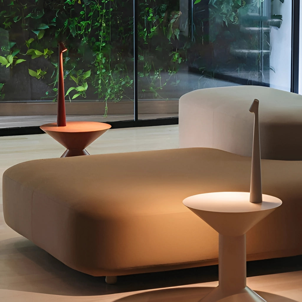 Portable rechargeable LED table lamp with aluminum construction, placed on a wooden table next to a green plant, featuring three light intensity settings, in a comfortable and stylish interior design setting.
