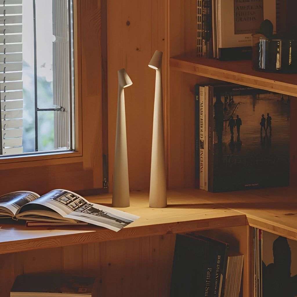 Portable rechargeable LED table lamp with aluminum construction, placed on a wooden writing desk near a window with shelves and books in the background.