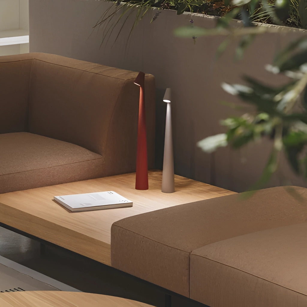 Portable rechargeable LED table lamp with aluminum construction on a wooden table, surrounded by a potted plant and a comfortable couch in a modern interior design setting.