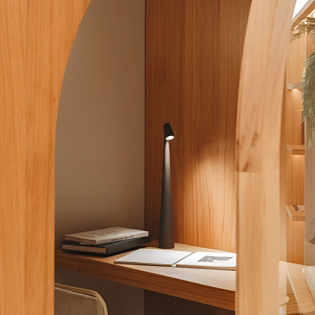 Portable rechargeable LED table lamp made of aluminum, placed on a wooden table with hardwood flooring in the background. The lamp offers three light intensities.