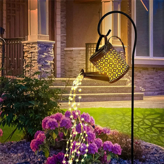 A solar watering can light with RGB/white LED hanging on a pole, illuminating flowers and plants in a garden with shades of blue and purple. The scene includes petals, grass, and groundcover, set in front of a window.