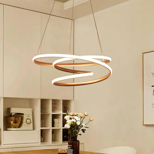 Elegant spiral pendant light with warm illumination hanging above a modern interior space featuring a wooden table with houseplants and a flower vase.