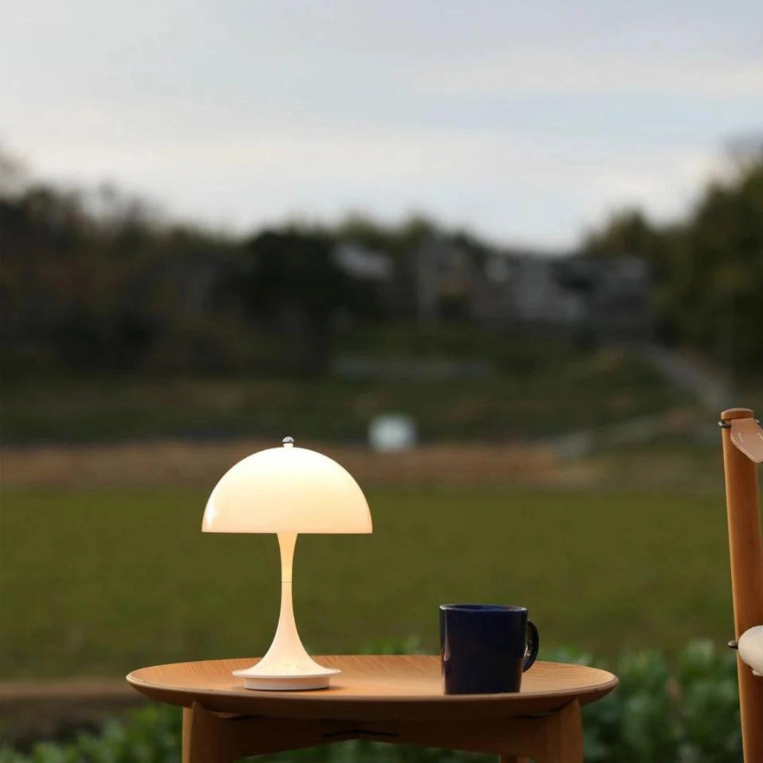 A group of Louis Poulsen Panthella 160 Portable Table Lamps in white, black, orange, gold, and gray, displayed outdoors on a wooden table with a grassy landscape and trees in the background.
