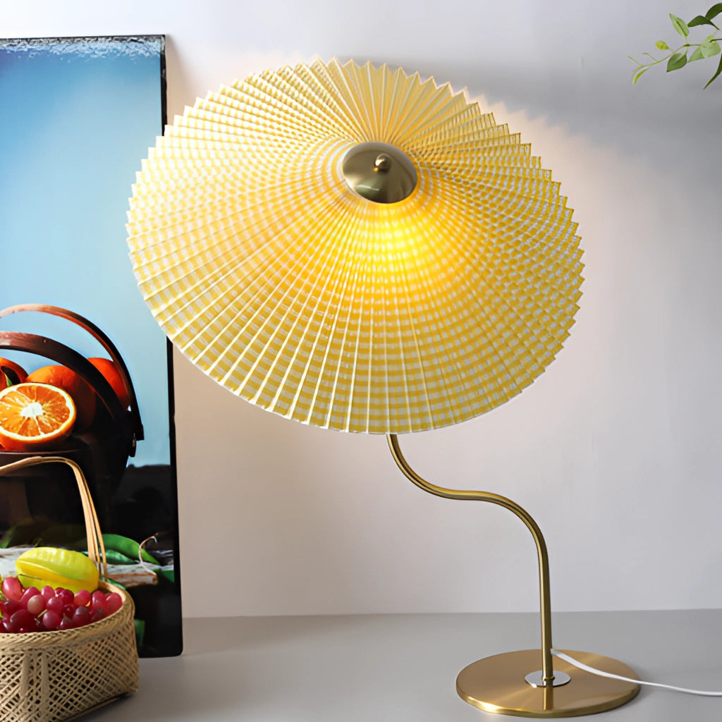 A table lamp with an umbrella-shaped shade featuring a yellow diamond, khaki, and white floral design, set against a dark green background. The lamp is illuminated, creating a warm, amber light, and is accompanied by a small potted plant.