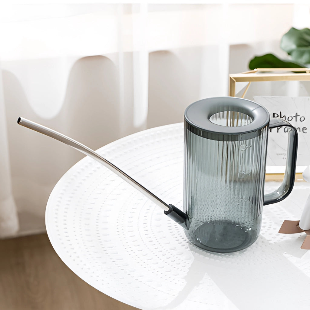 Grey watering can with extended spout on a table next to drinkware and dishware.
