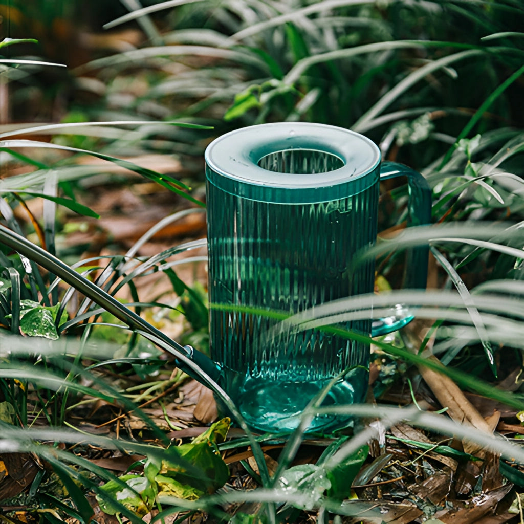 Watering can with extended spout being used to water plants in a lush, green garden with grass and various types of vegetation.