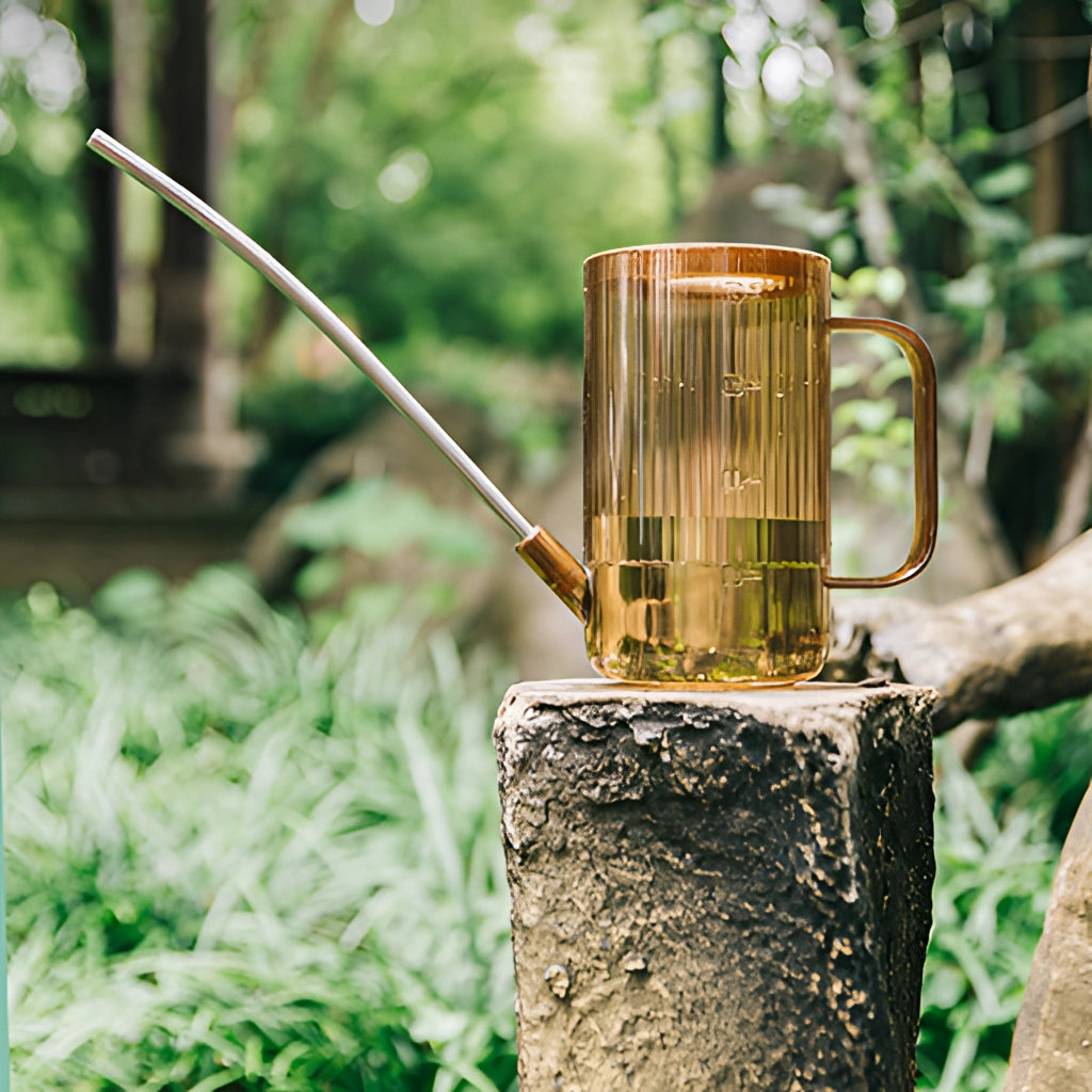 A green watering can with an extended spout positioned near a tree in a garden with grass and wooden elements in the background.