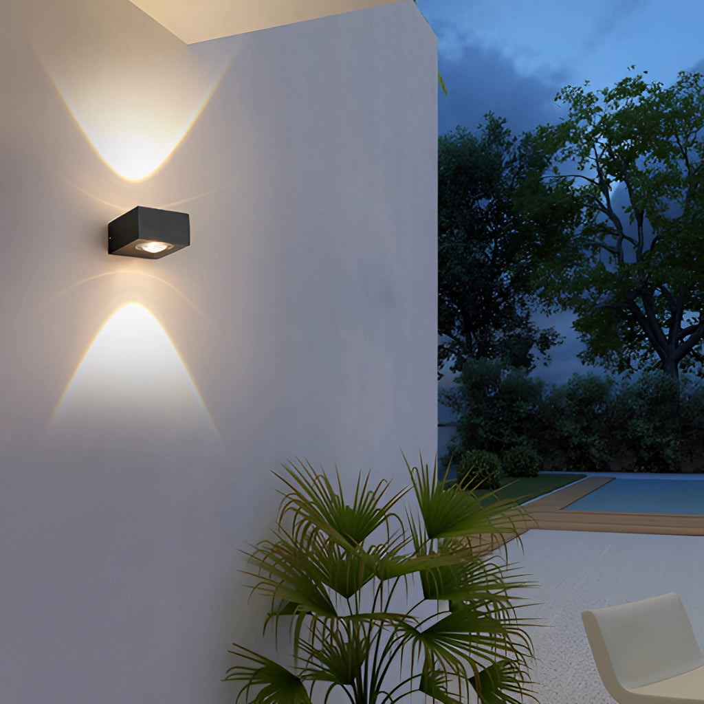 Black UFO-shaped waterproof wall light mounted outdoors with a glowing light, surrounded by plants and trees against a clear sky.