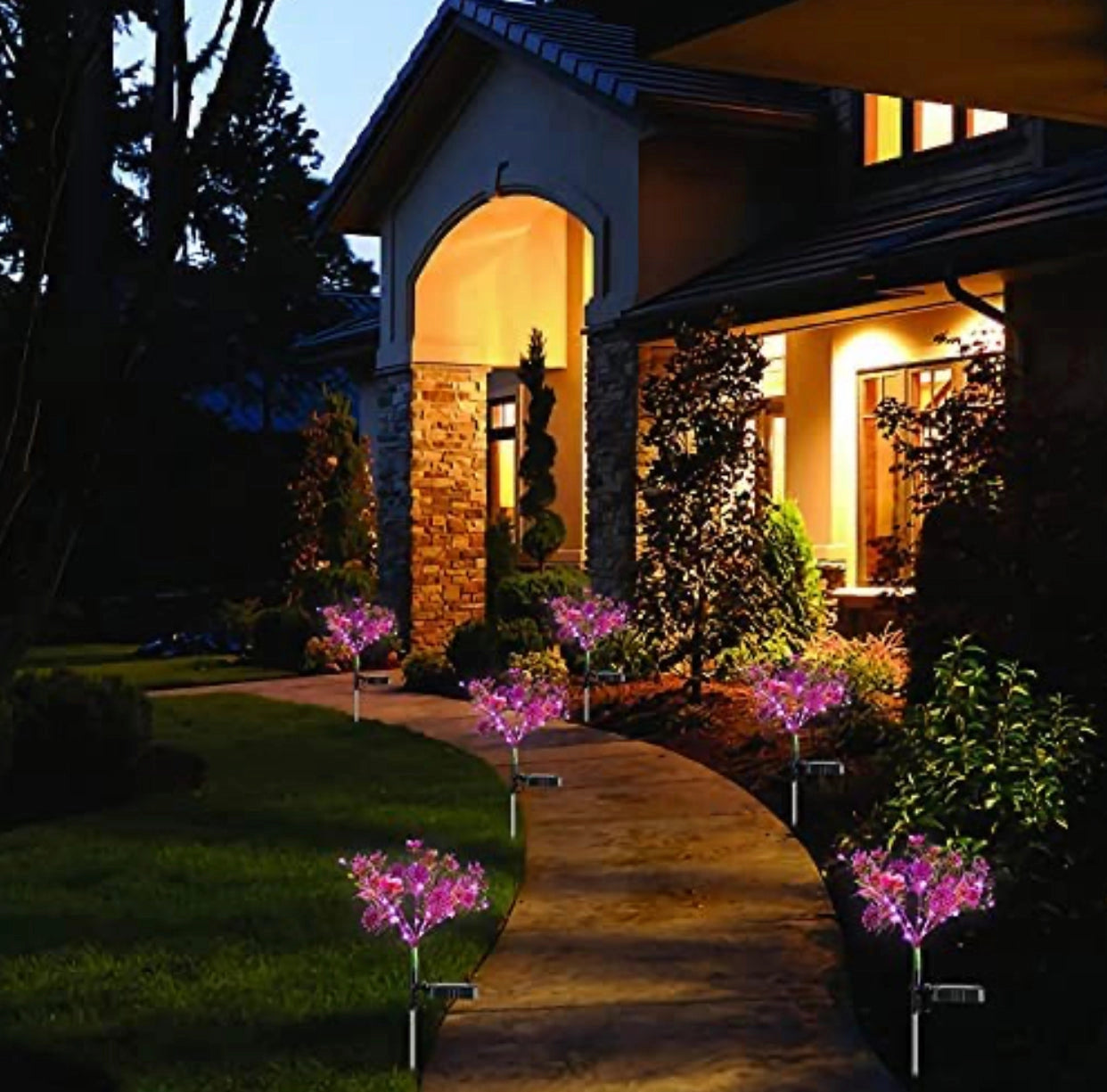 Purple Waterproof Solar Phalaenopsis LED Flower Lights illuminating a garden, with a house and trees in the background under a clear sky.